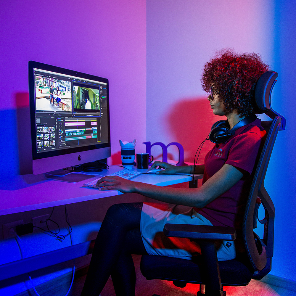 A woman with curly red hair sitting in a supportive computer chair in front of a mac on a desk.